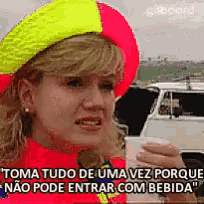 a woman wearing a neon yellow and red hat is holding a cup and says " toma tudo de uma vez porque "