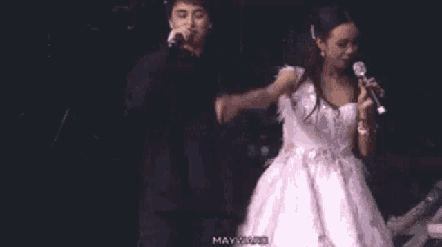 a man is singing into a microphone while a woman in a white dress stands next to him on a stage .
