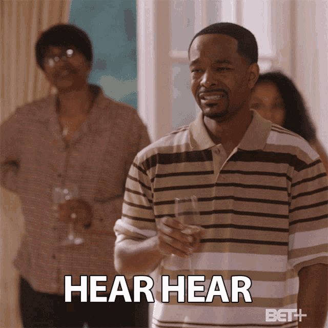 a man in a striped shirt is holding a glass of wine and the word hear hear is on the bottom
