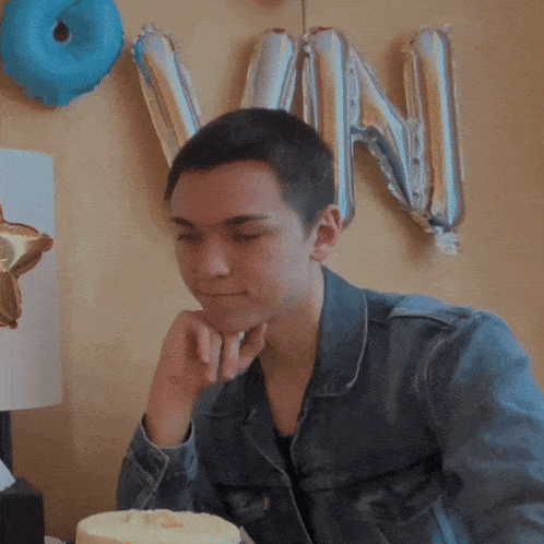 a young man in a denim jacket looks at a cake in front of balloons that spell out the letter vn