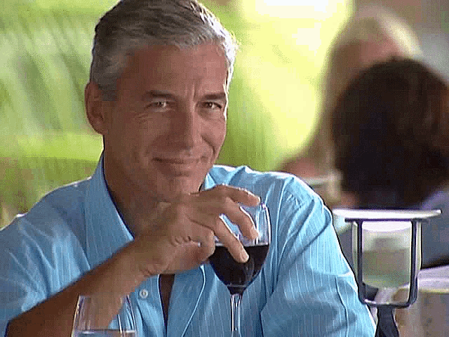 a man in a blue shirt is holding a glass of wine and smiling