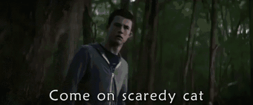 a man and a woman are standing in the woods with the words come on scaredy cat written on the screen .