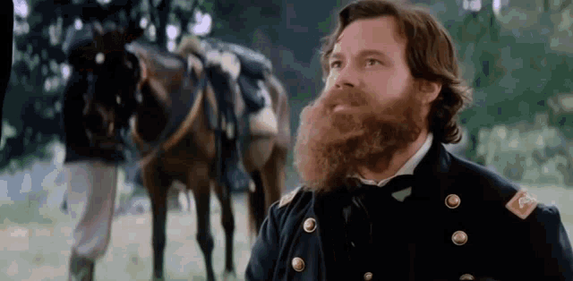 a man with a beard is standing in a field with a horse in the background .