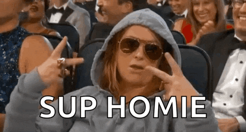 a woman wearing a hoodie and sunglasses is sitting in a crowd of people and making a funny face .