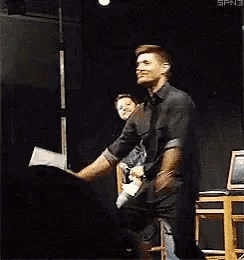 a man in a black shirt is dancing on a stage while another man sits in the background .
