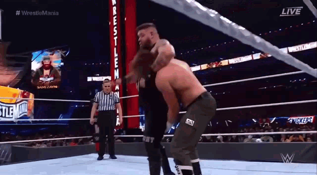 two men are wrestling in a wrestling ring with a referee and a snickers sign in the background