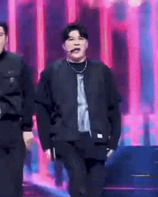 a man in a black jacket is dancing on a stage with a pink background .