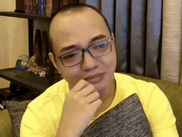 a man wearing glasses and a yellow shirt is sitting on a couch with a pillow .