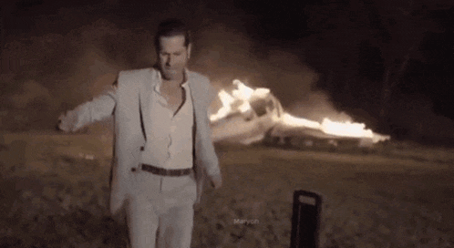 a man in a suit and suspenders is standing in front of a burning car .