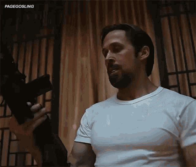 a man in a white t-shirt is holding a gun in front of a curtain .