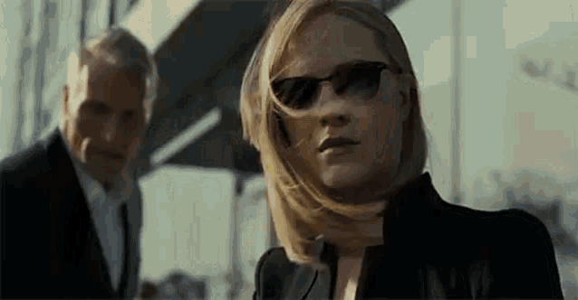 a woman wearing sunglasses is standing in front of a man in a suit and tie .