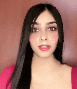 a woman with long black hair and pink cheeks is wearing a pink shirt and looking at the camera .
