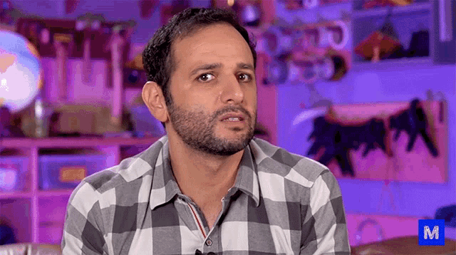 a man in a plaid shirt is sitting in front of a purple wall with the letter m in the corner