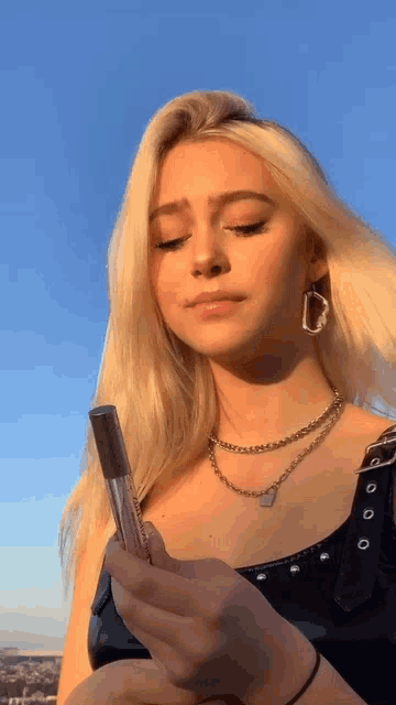 a blonde woman wearing a necklace and earrings is holding a lip gloss in her hand .