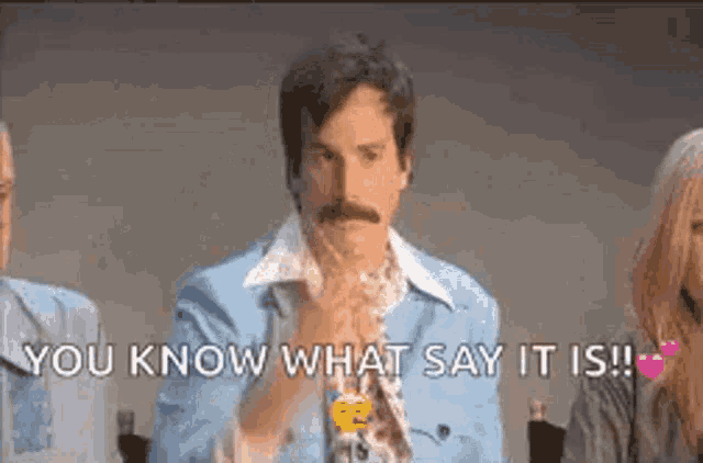 a man with a mustache is sitting in front of a group of people and says `` you know what say it is ! ''