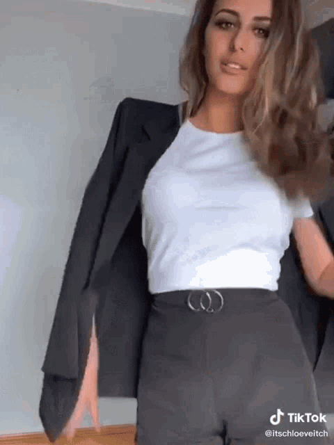 a woman in a white t-shirt and black pants is holding a blazer over her shoulder .