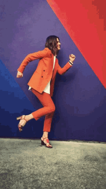 a woman in an orange suit and heels is running in front of a colorful wall