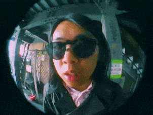 a woman wearing sunglasses looks through a fisheye lens at the camera