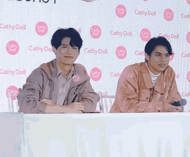 two men are sitting at a table with a microphone in front of a wall that says cathy doll