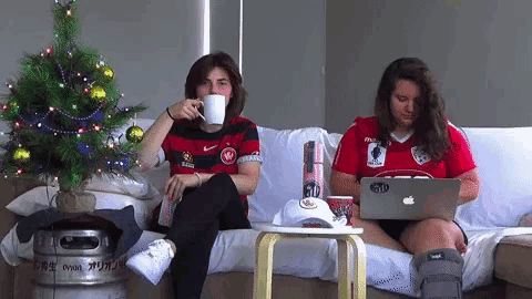 two women sit on a couch with one using an apple laptop and the other drinking from a mug