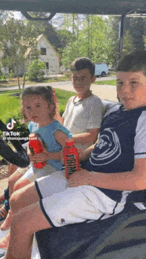 three children are sitting in a golf cart and one of them is holding a bottle of primal energy drink