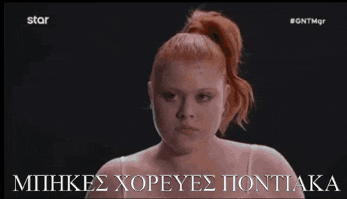 a woman with red hair and a ponytail looks at the camera with a caption in greek