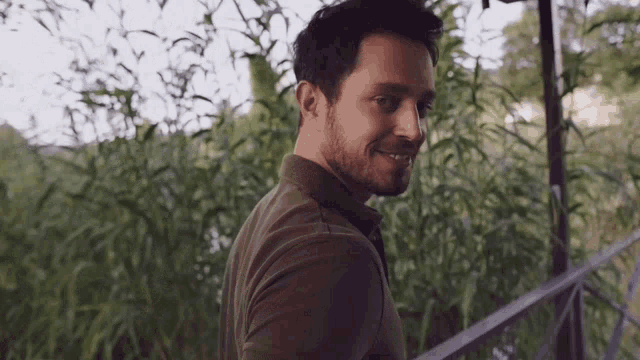 a man with a beard wearing a green shirt is smiling in front of tall grass