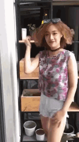 a woman in a floral top and white shorts is standing in front of a shelf with potted plants on it