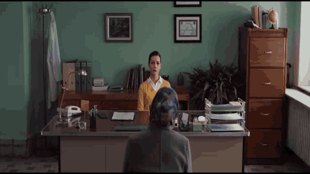 a woman in a yellow sweater sits at a desk in front of a green wall