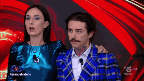 a man in a blue plaid jacket stands next to a woman in a blue top with the hashtag grandefratello on the bottom