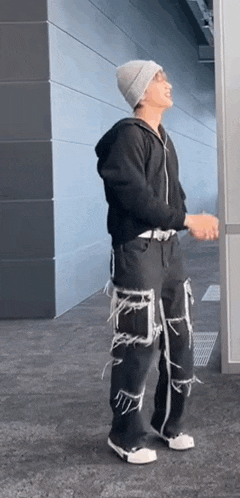 a man wearing a beanie and a black hoodie is standing next to a wall .