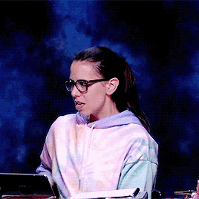 a woman wearing glasses and a tie dye hoodie sits at a desk