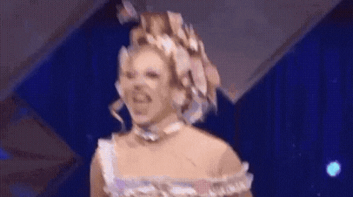 a drag queen wearing a white dress and a flower crown is dancing on a stage .