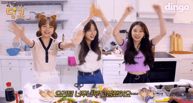 three girls are standing in a kitchen with their arms in the air and the word dingo in the corner