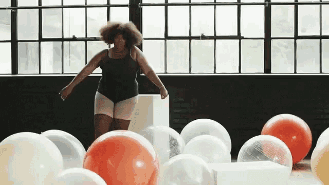 a woman in a black tank top stands in a room surrounded by balloons