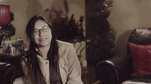 a woman wearing glasses and a tan jacket is laughing in a living room
