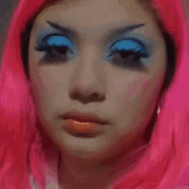 a woman with pink hair and blue eye makeup looks at the camera .