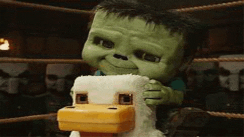 a green stuffed animal is sitting on top of a stuffed chicken in a boxing ring .