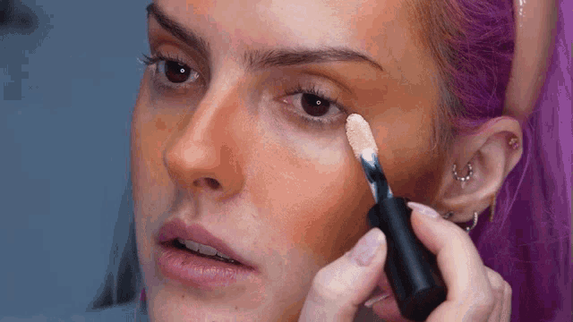 a woman with purple hair is applying concealer to her face .