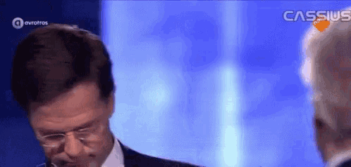 a man in a suit and tie is talking to another man in front of a blue background .