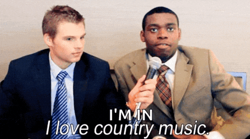 a man in a suit and tie is holding a microphone while another man says i 'm in i love country music