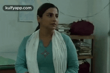 a woman in a blue shirt and white scarf is standing in a room with a stack of books on a shelf .