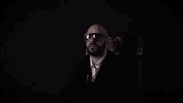 a man with a beard is sitting in a chair wearing sunglasses and a suit .