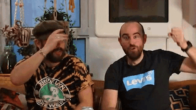 two men are sitting on a couch with their arms in the air . one of the men is wearing a levis shirt .