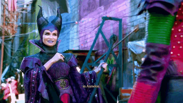 a woman in a purple costume with horns is holding a wand and says " in auradon "