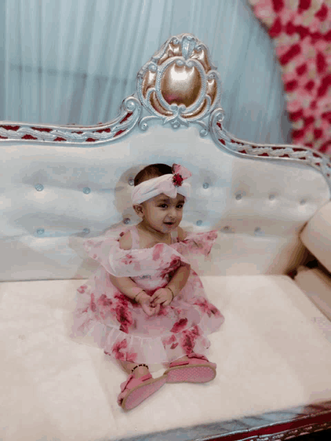 a baby girl in a pink dress and headband sits on a white couch