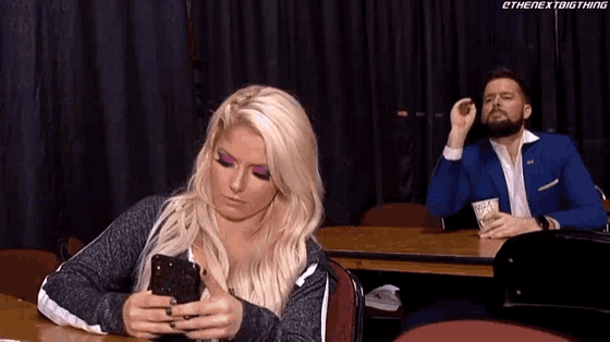 a woman is sitting at a table looking at her phone while a man in a blue suit looks on .