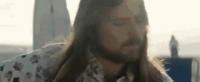 a man with long hair and a beard is wearing a floral shirt and looking down .
