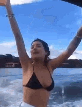 a woman in a black bikini is standing on a boat with her arms in the air .