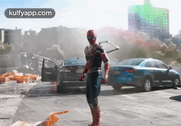 a man in a spiderman costume is standing in a parking lot next to cars .